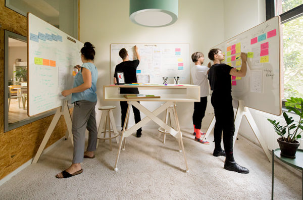 Vier Menschen stehen ein einem Raum und schreiben an Designwhiteboards. In der Mitte steht ein Stehtisch auf Rollen