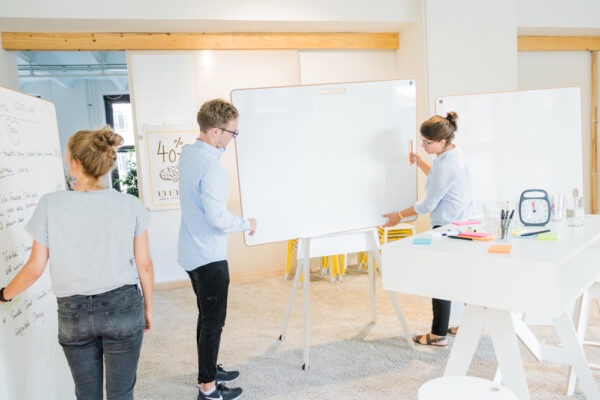 Zwei Menschen in einer Workshopsituation verschieben ein Whiteboard im rollbaren Gestell